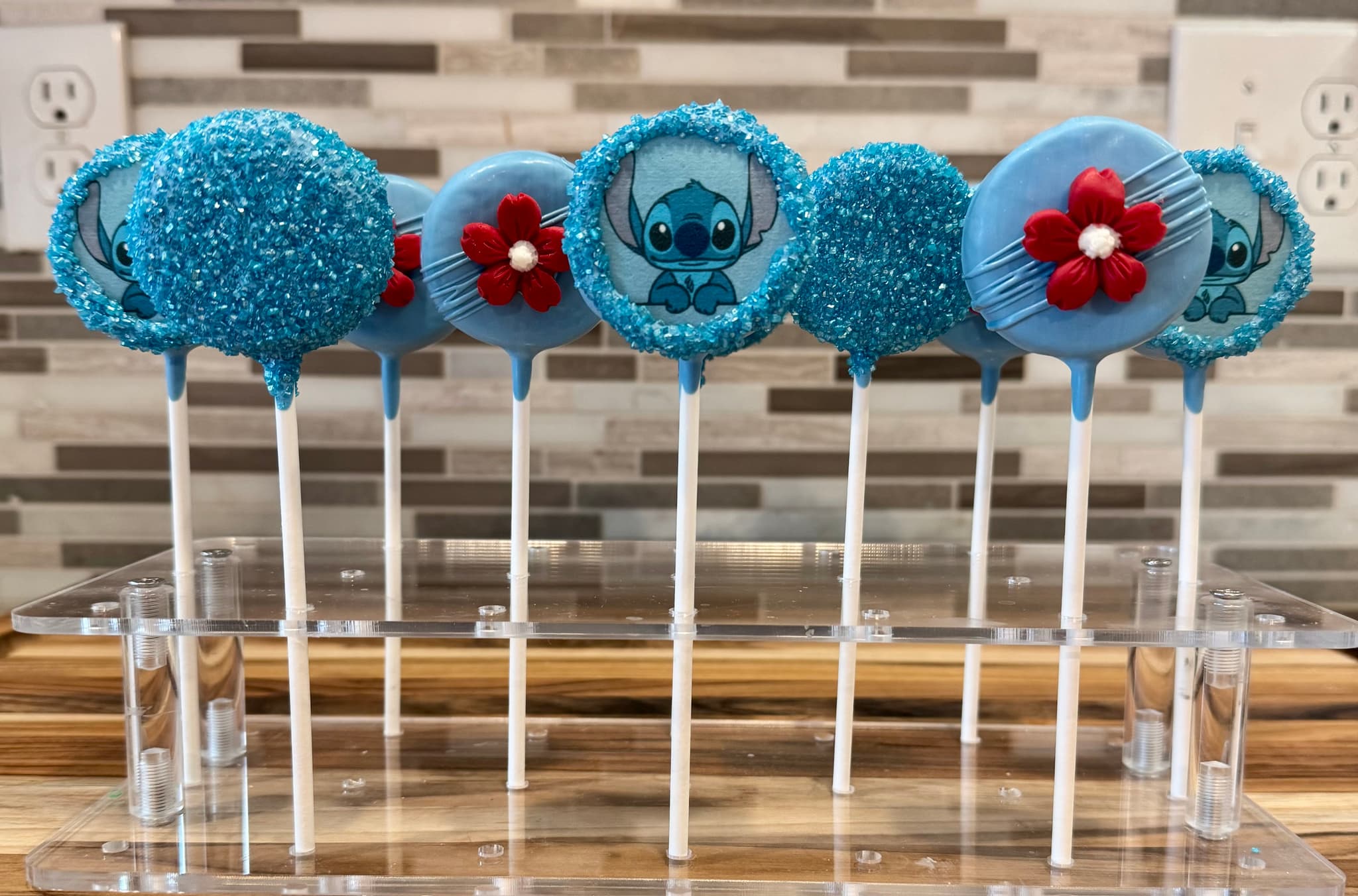 Blue cake pops with a printed Stitch character design, red fondant flowers, and some rolled in blue sparkling sugar.