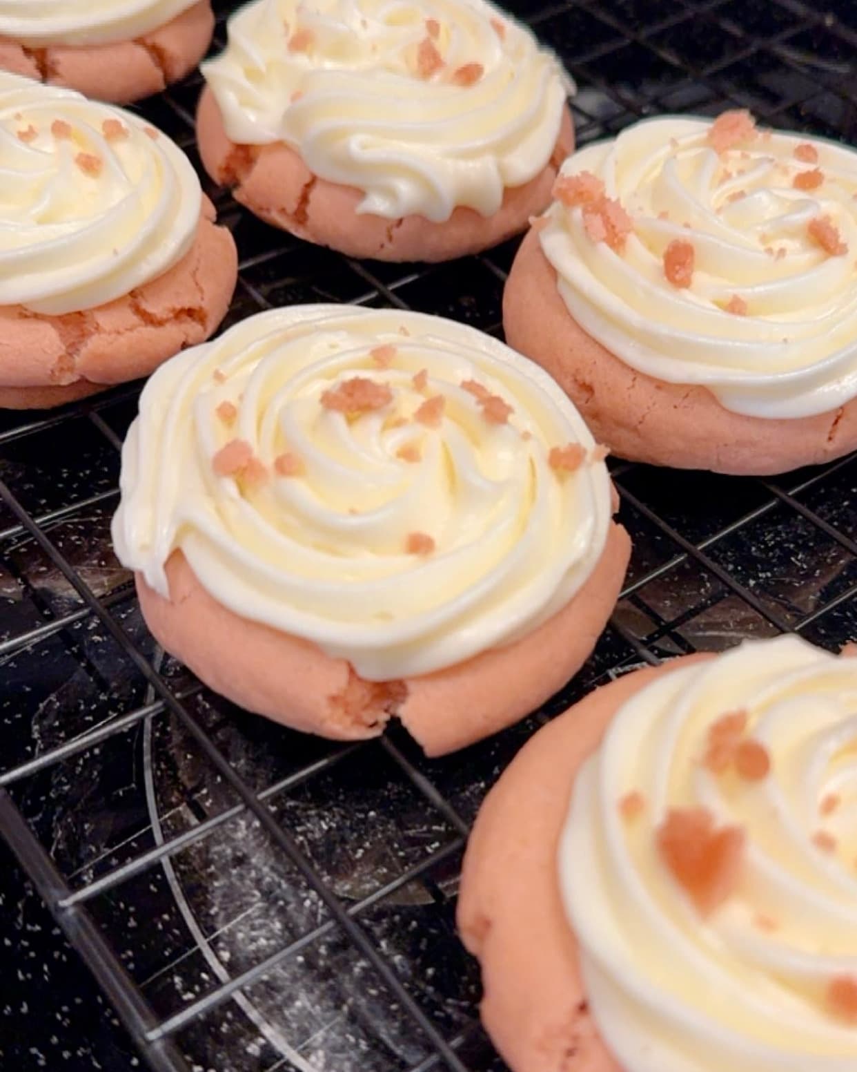 Frosted pink cookies with a swirled cream cheese frosting top and orange sugar crystals, cooling on a wire rack.