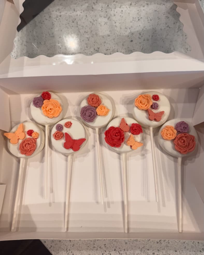 White chocolate cake pops decorated with orange, red and purple fondant roses and butterflies, in a bakery box.