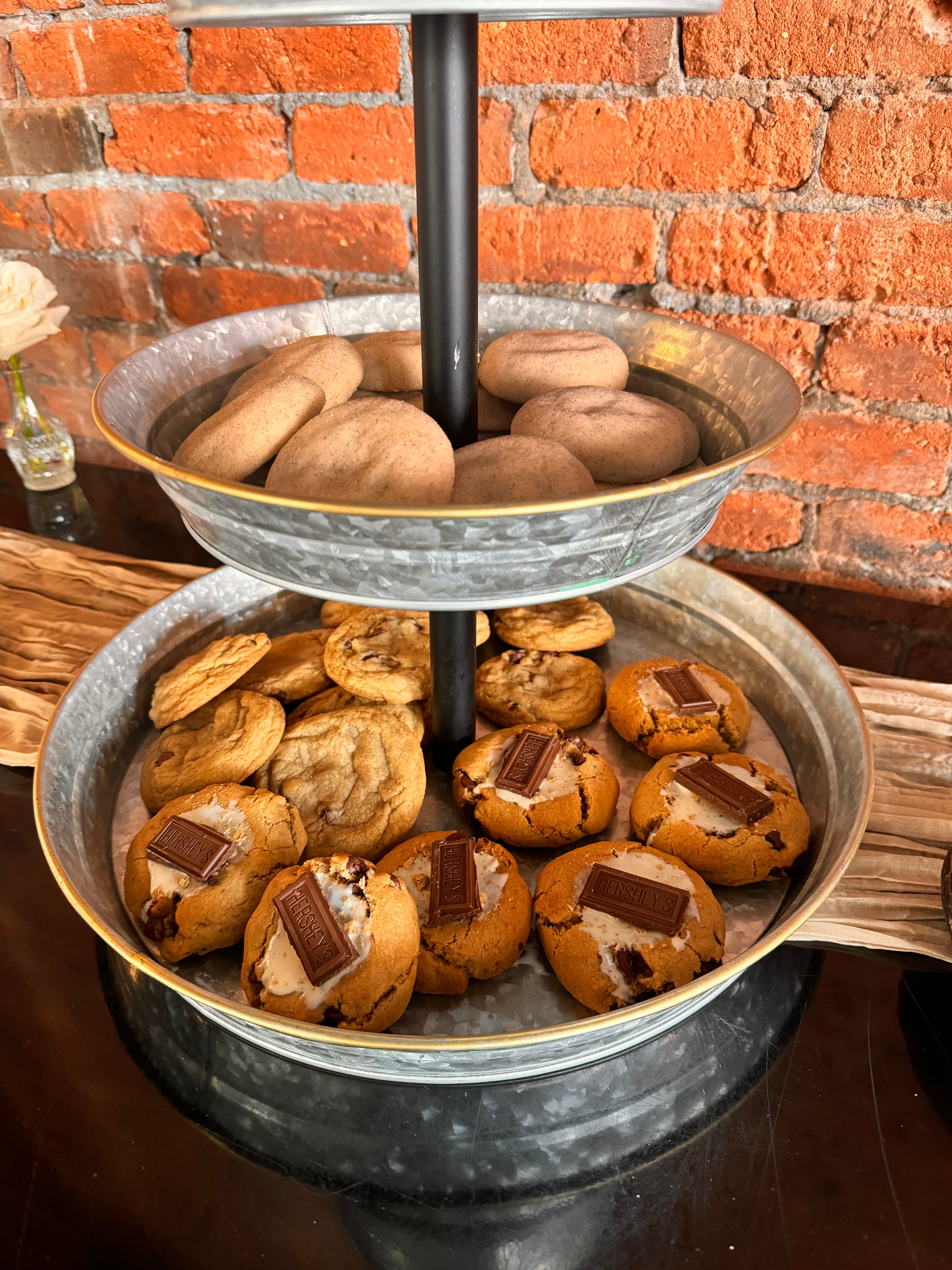 Chocolate chip cookies topped with chocolate squares, and snickerdoodles, piled in two galvanized tins on a stand against a brick wall.