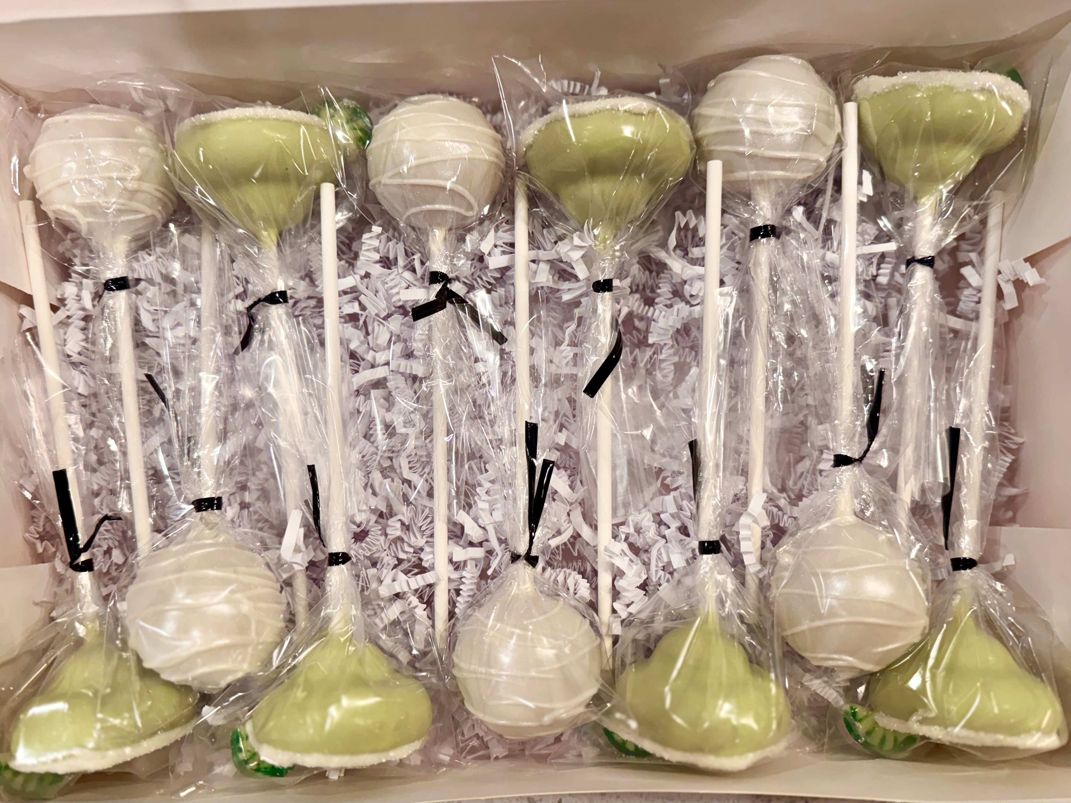 Sage green and white cake pops wrapped in cellophane with black ribbon ties, nested in white paper shred in a gift box.
