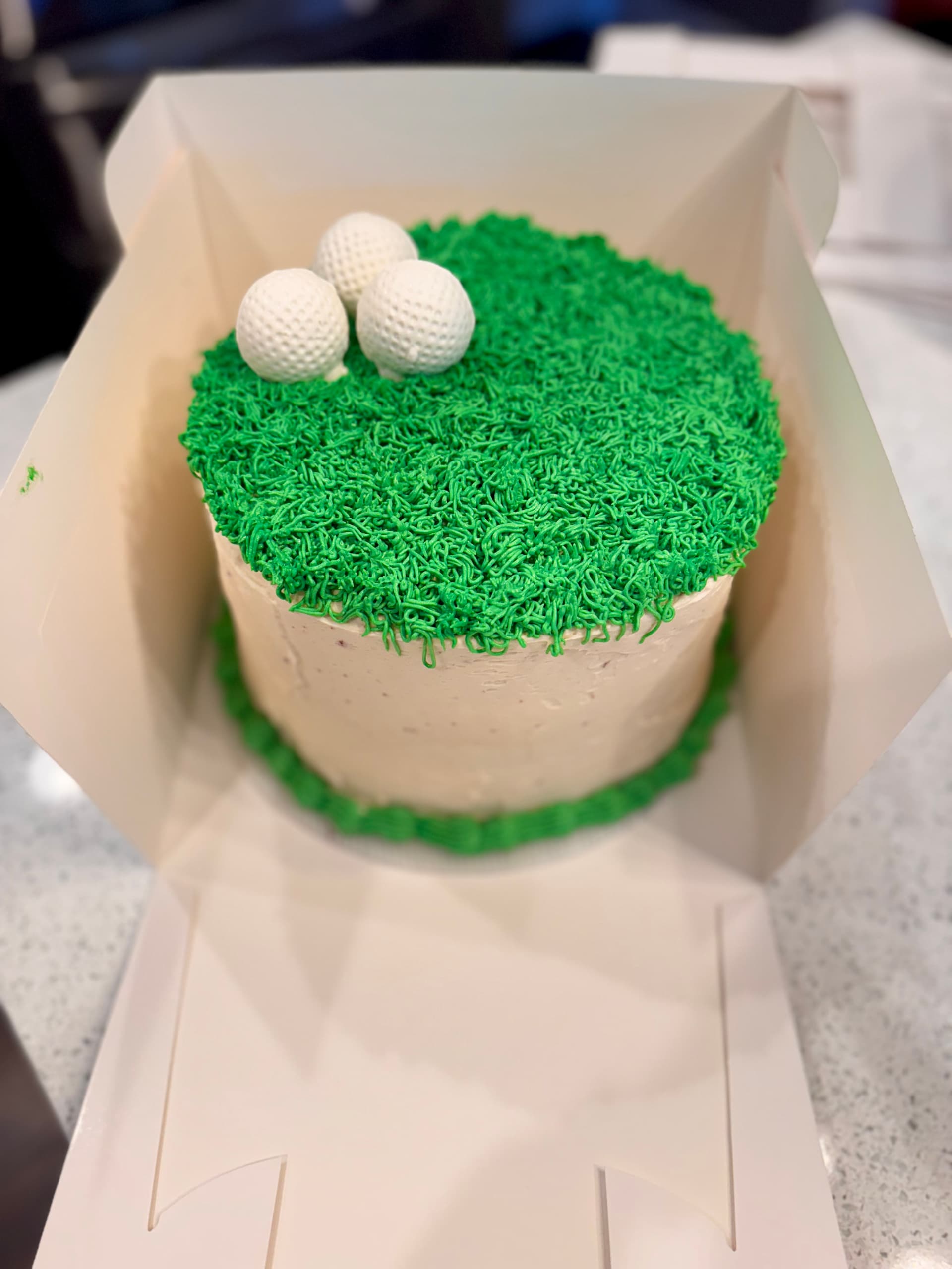 Round cake with green buttercream grass piping and fondant golf balls, boxed.