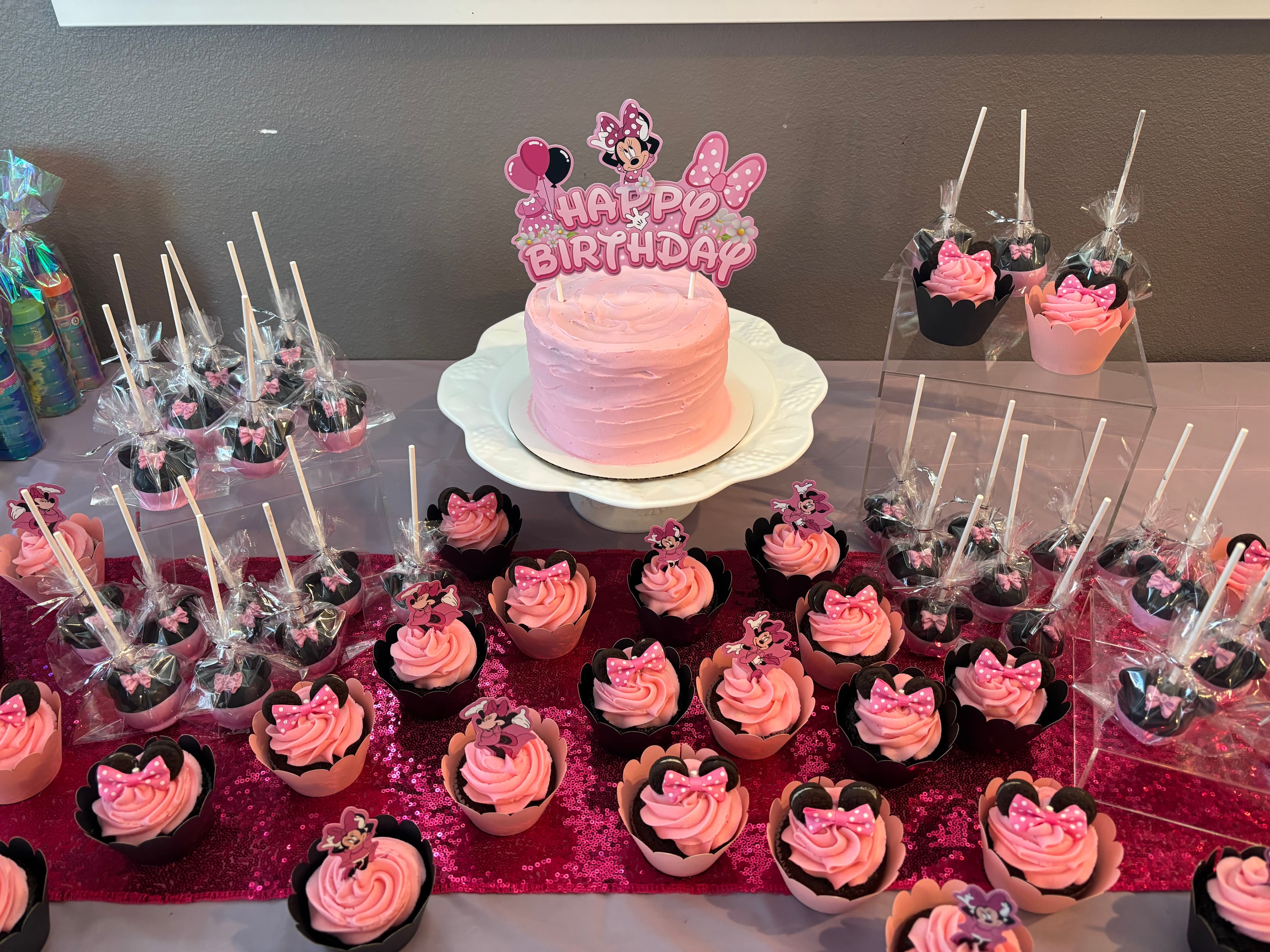 Birthday dessert table with a pink cake, pink-frosted cupcakes and wrapped cake pops on a pink sequin runner, under a Minnie Mouse happy birthday topper.