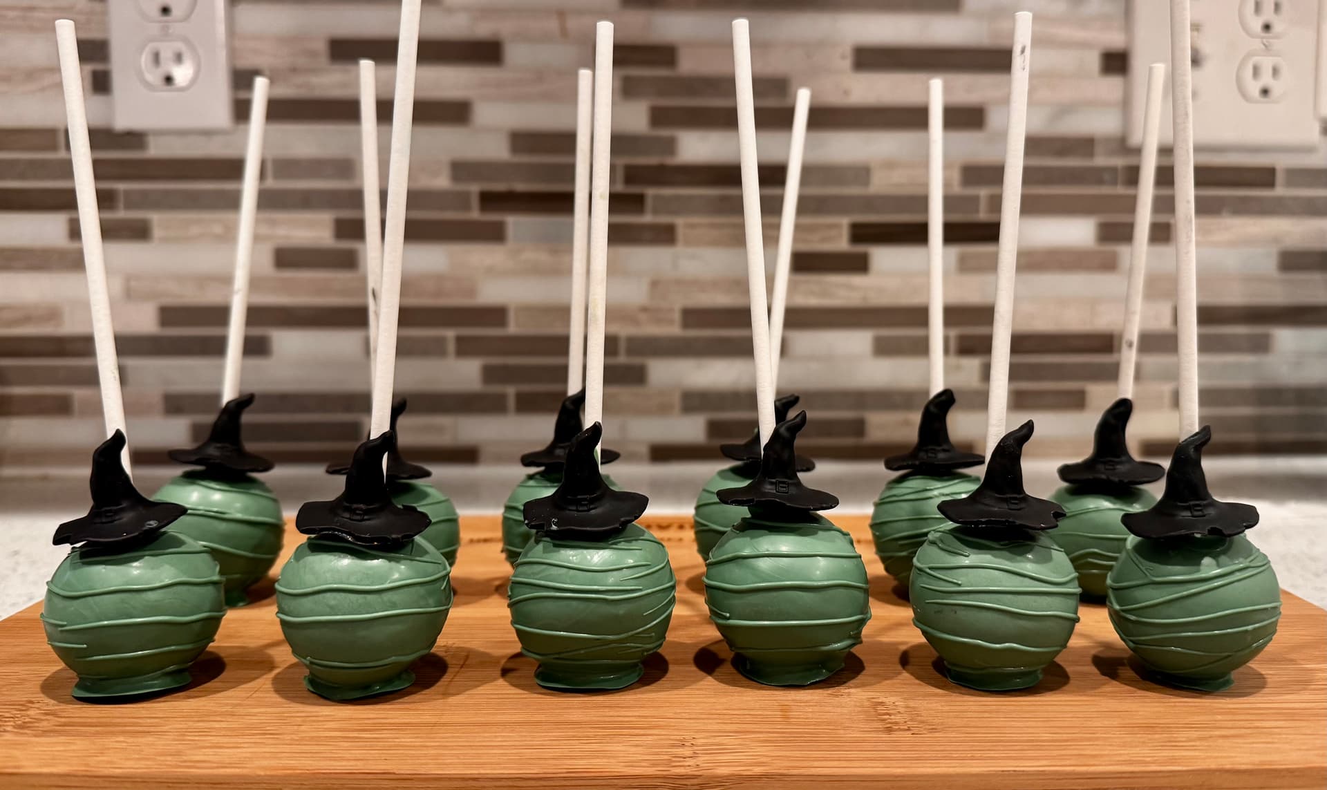 Pearlescent pink cake pops, each topped with a small white fondant crown, lined up on a wood board.