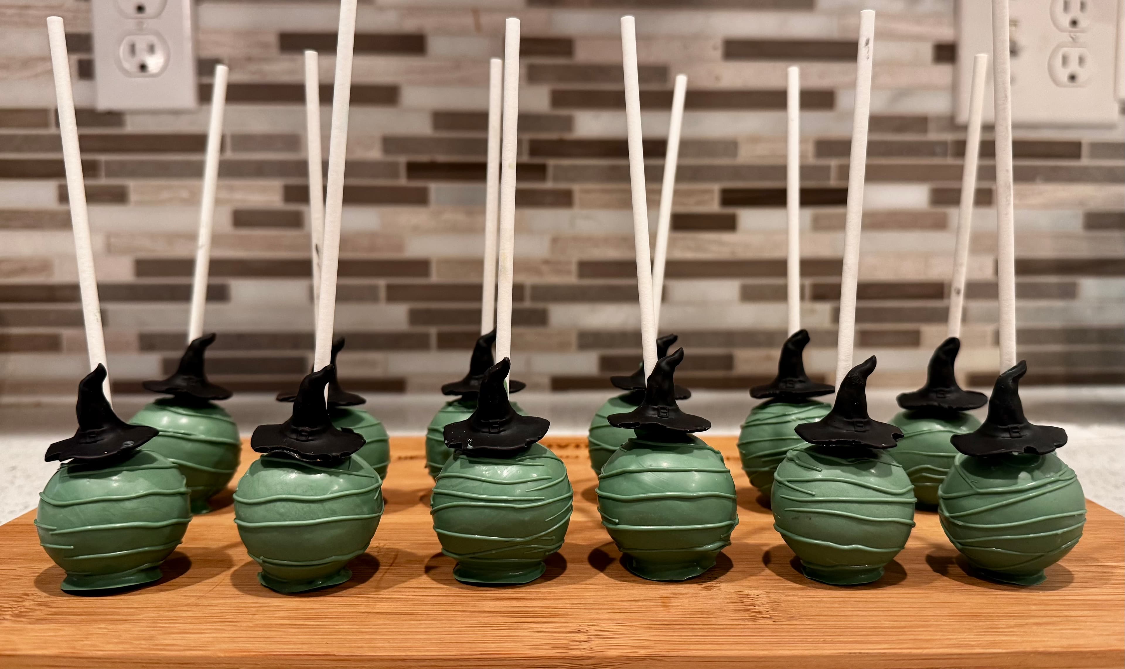 Dark green cake pops, each topped with a black witch hat, lined up on a wood board.