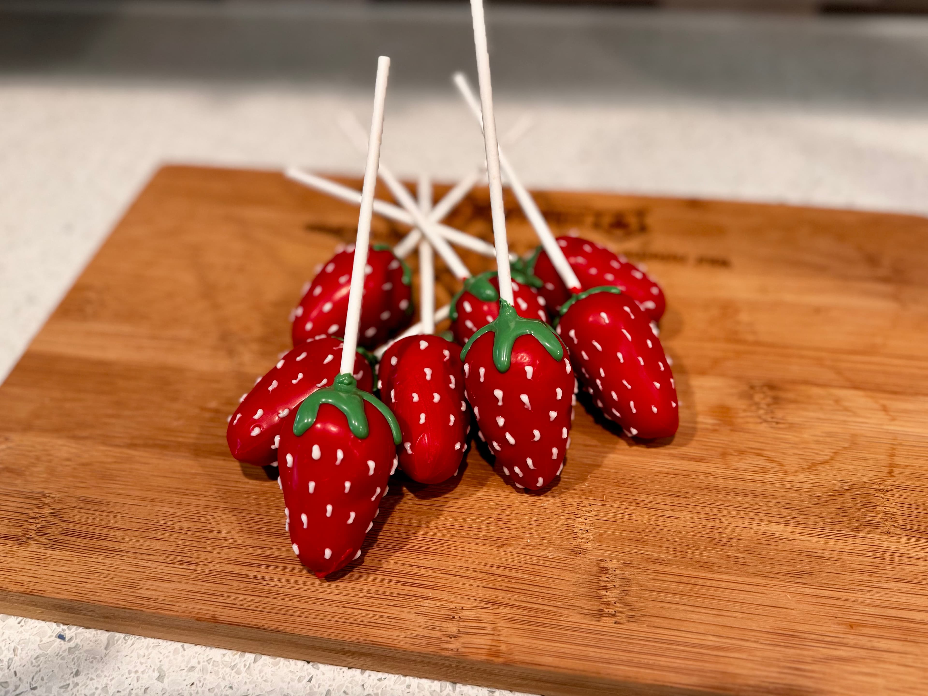 Strawberry-shaped red cake pops with white seed detailing and green tops, clustered on a wood board.