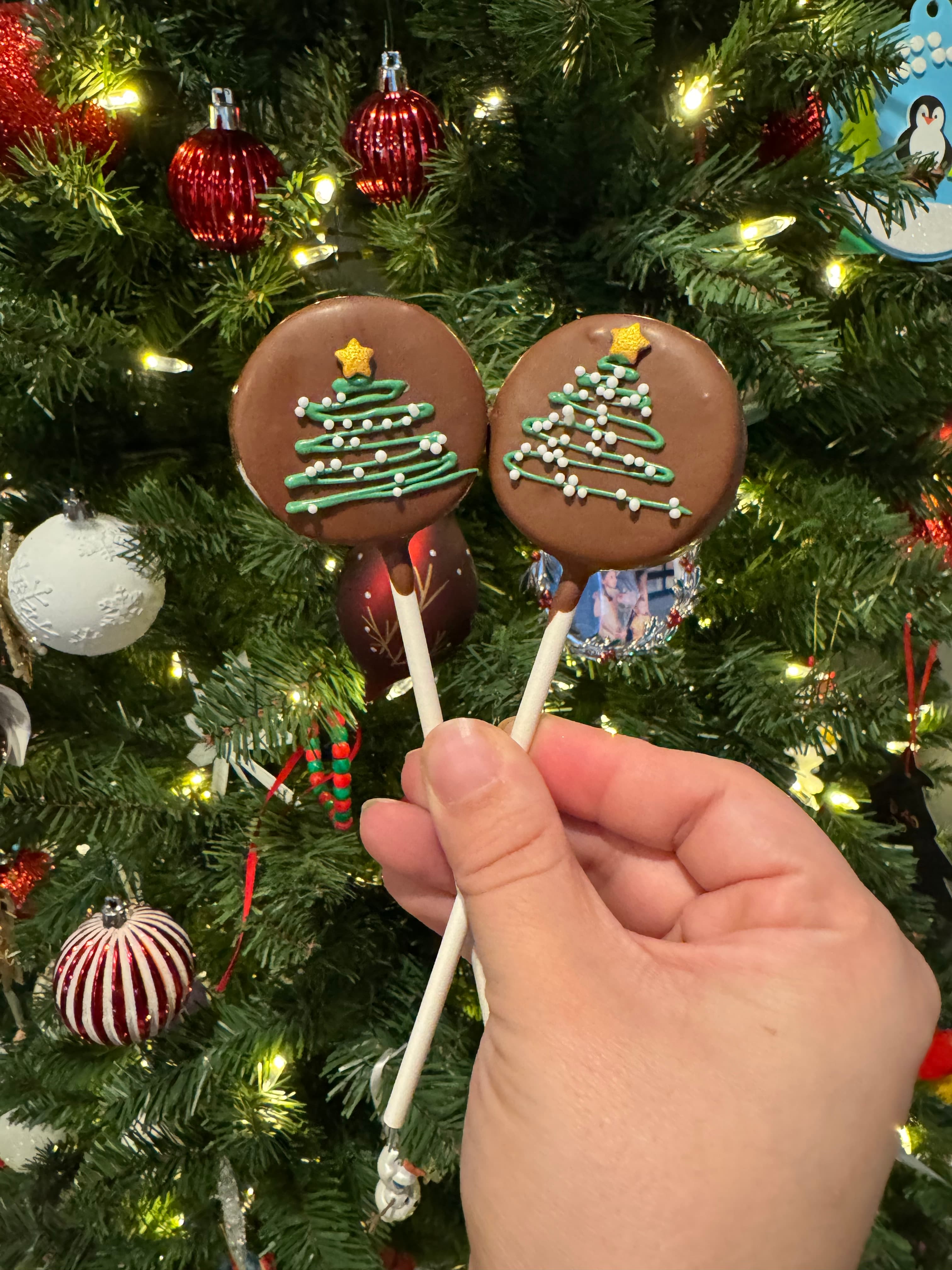 Two round chocolate cookies piped with a green Christmas tree, held in front of a Christmas tree.