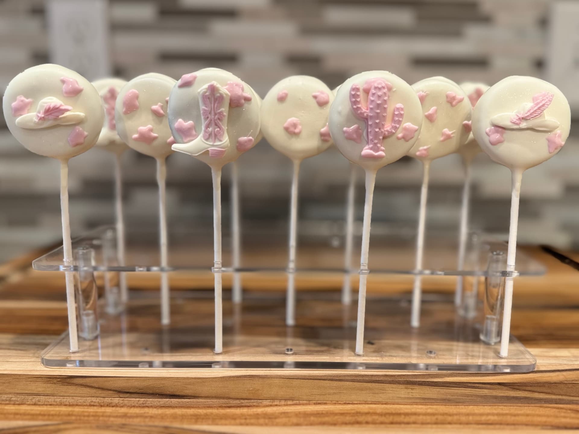 Cream cake pops decorated with a pink cowboy hat, boot and cactus, on a stand.