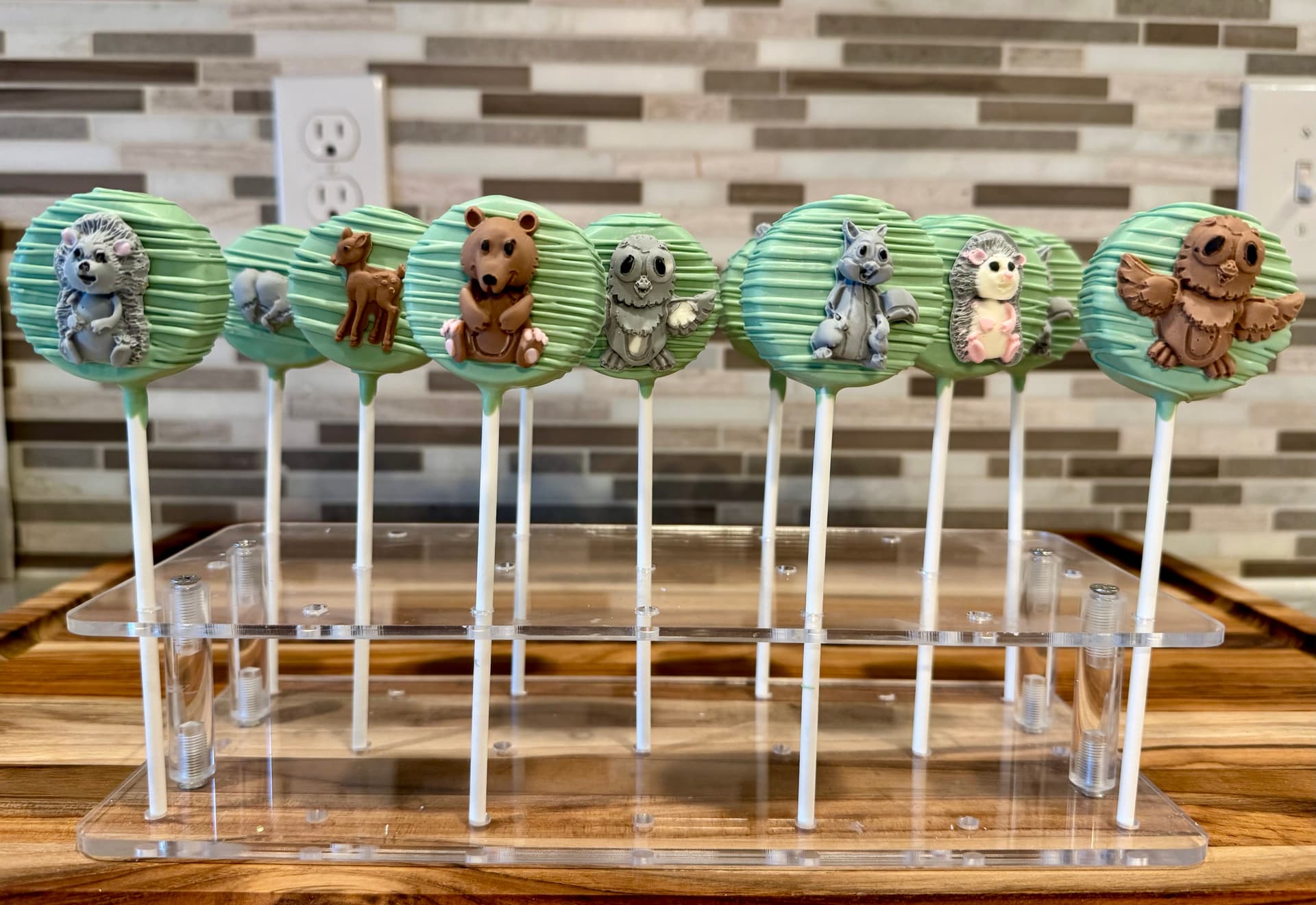 Woodland-animal cake pops in pale blue, with a bear, raccoon, hedgehog and owl among painted trees, on a stand.