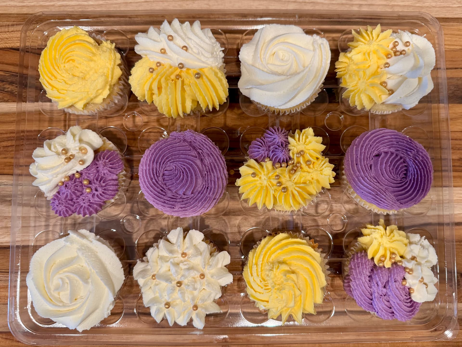 A dozen cupcakes with swirled buttercream in yellow, white and purple, some topped with gold pearl sprinkles, in a bakery tray.