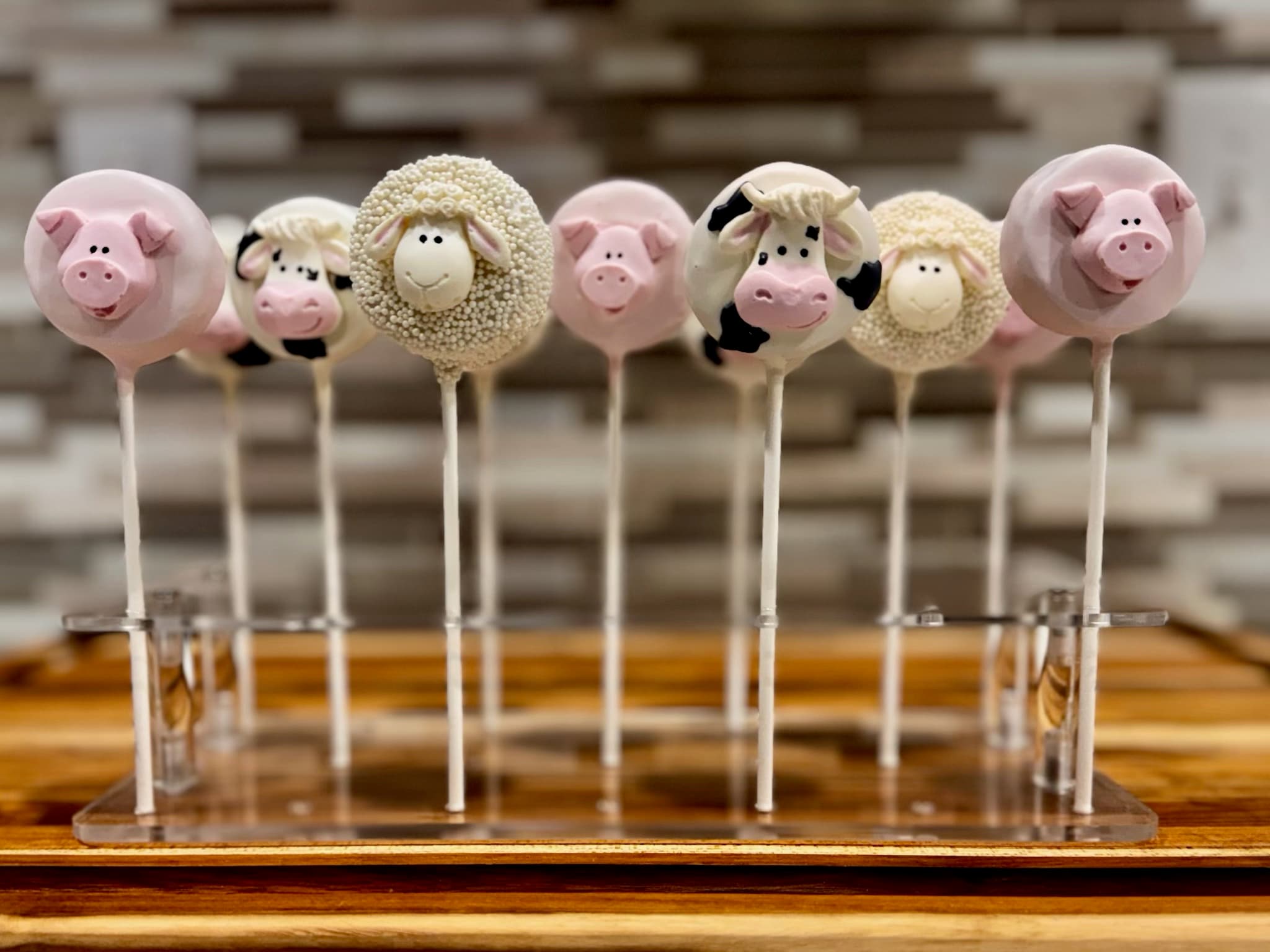 Farm animal cake pops with pink pig faces, black-and-white cow faces and cream sheep covered in white nonpareils, on a clear stand.